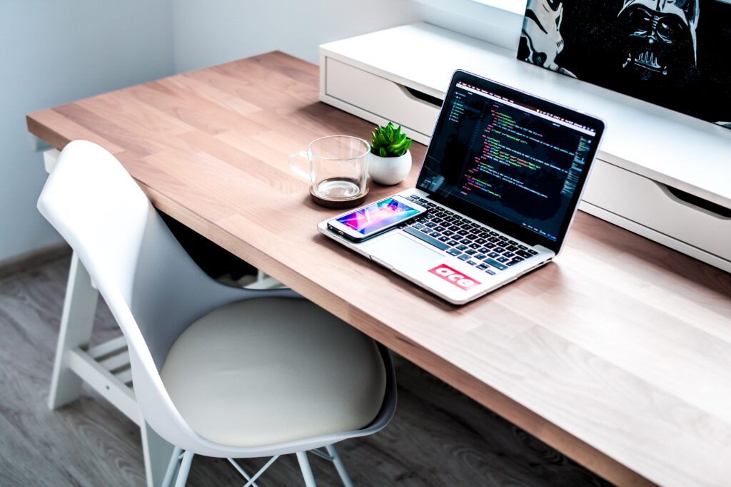 Laptop displaying code on a wooden desk beside a smartphone, glass mug, and a small succulent