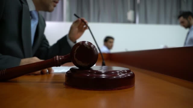 Gavel resting on a sound block in a courtroom, with people seated at a conference table behind