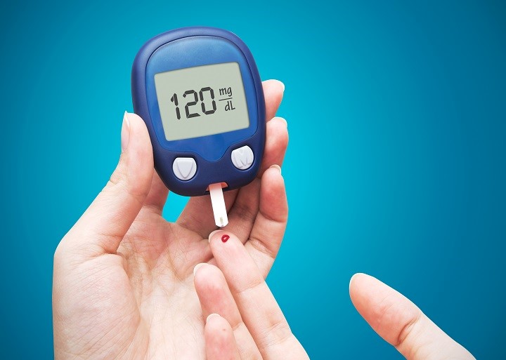 Hand holding a blood glucose meter reading 120 mg/dL next to a pricked fingertip